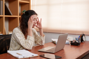 Stressed Asian woman at desk pinches bridge of nose, gesture of tension from prolonged computer....