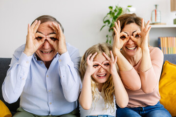 Portrait of happy multi-generational family having fun doing funny faces with fingers sitting together on sofa at home. Cheerful headshot of grandfather, mother and small daugther