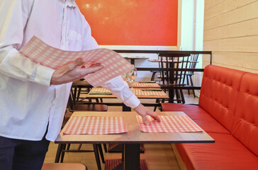 Unrecognizable man laying tablecloths for dishes on a restaurant table, preparing the dining room