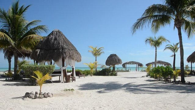 HD video of a tropical public resort with thatched huts and palm trees, clear blue skies, white sands with calm blue waters, and sunny skies. Holbox Island, Mexico