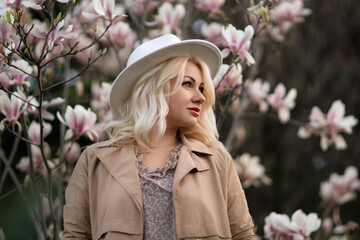 A blonde woman wearing a white hat stands in front of a tree with pink flowers