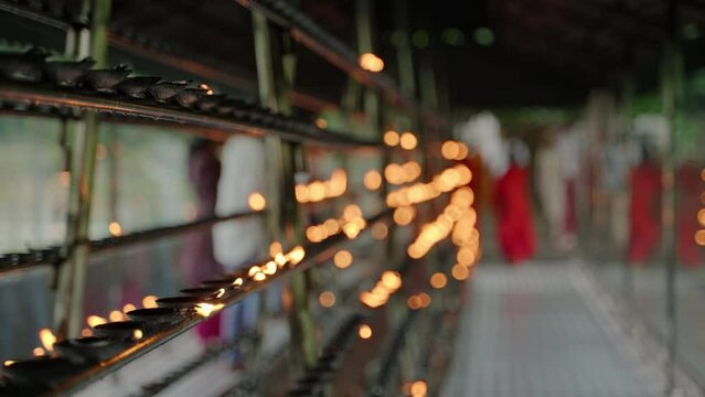 Buddhist devotees light oil lamps, offer prayers in temple. Rows of flickering flames line racks with blurred figures moving, serene religious practice in sacred place, creating peaceful atmosphere.