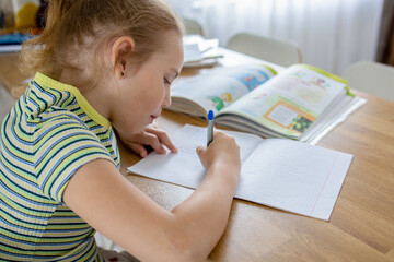 A charming little girl does her homework based on a textbook and transfers the information to a workbook. The idea of integrating online and home learning.