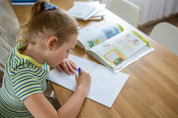 An image of a lovely blonde baby who is doing her homework, using textbooks and a workbook. The concept of distance learning online and at home.