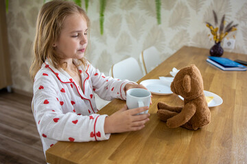 A cute blonde girl in beautiful pajamas with hearts is having a delicious breakfast and feeding her...