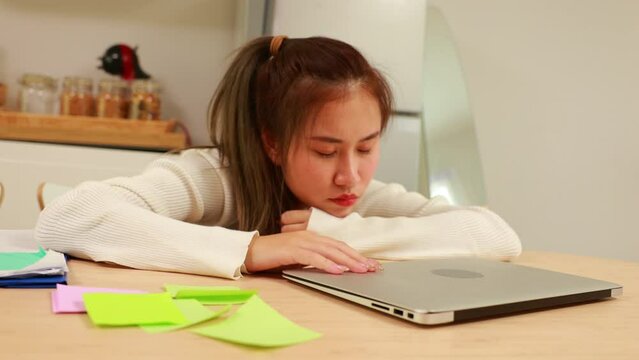 Tired exhausted, fatigue and fell asleep. Yaoung asian thai female working on laptop in kitchen room at home. Overwork. Exhausted woman  sleeply eye at workplace