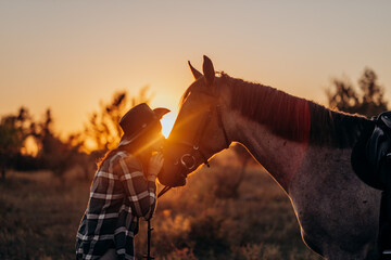 Beautiful smiling young woman in hat and shirt playing with brown horse in a field at sunset....