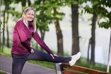 Active middle-aged woman stretches leg on park bench, surrounded by lush greenery