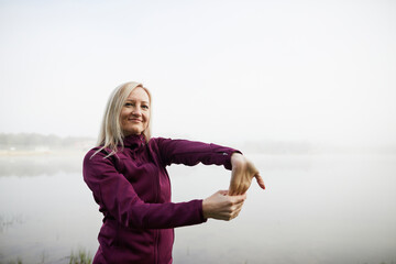 Confident woman stretching by a misty lake, with a serene expression and a background blurred by fog