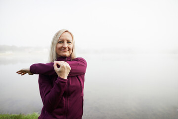 Mature woman stretches her arms while enjoying a misty morning by a serene lake