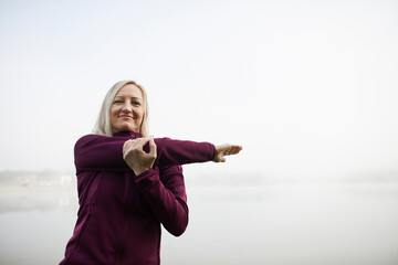 Mature woman practices stretching exercises by a foggy lake, emanating serenity and focus