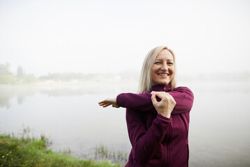 A woman in her forties stretches with a smile, by a misty lakeside, wearing a purple jacket