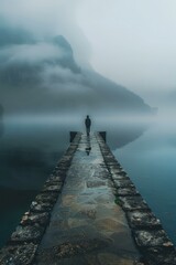 Man standing alone on a pier in the middle of a foggy lake with mountains in the distance