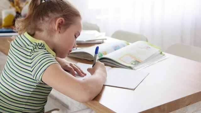 A cute blonde school-age girl is doing her homework sitting at the kitchen table. The child does written assignments in a workbook. The concept of online and home education.