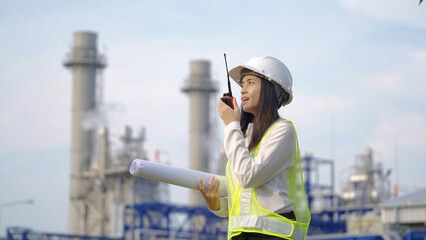 A female engineer in a hard hat and reflective vest holds blueprints and communicates via walkie-talkie at an industrial power plant.
