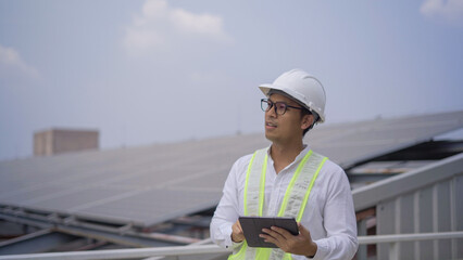 An engineer in safety gear uses a digital tablet to inspect and manage solar panels installed on the roof of an industrial building.
