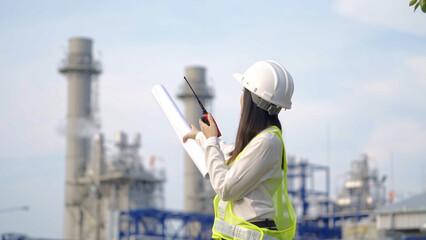 A female engineer in a hard hat and reflective vest holds blueprints and communicates via walkie-talkie at an industrial power plant.

