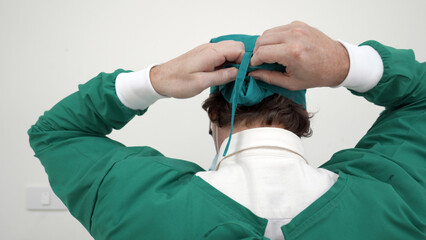 A surgeon in green scrubs meticulously ties his surgical cap at the back, preparing for a medical procedure in a clinical environment.
