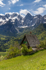 An idilic hut by the field of grass in front of a mighty mountain range in the background.