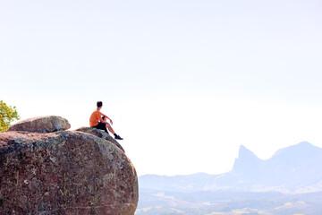 A man sitting on the edge of a cliff, gazing at mountains in the background. Ideal for outdoor enthusiasts such as hikers or those seeking personal growth through travel