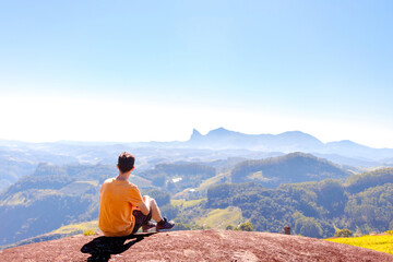 A Brazilian man in a yellow t-shirt rests atop a mountain, gazing at lush green hills under a sunny blue sky. Seeking stress relief or happiness through nature immersion