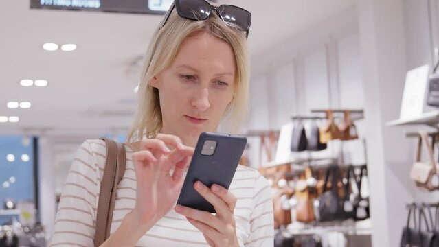 Beautiful female customer shopping in clothing store using smartphone, browsing online, comparing online, choosing stylish clothes. Fashion store, colorful brands,