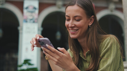 Woman with brown hair, dressed in an olive green sweater, using mobile phone app texting sms on...