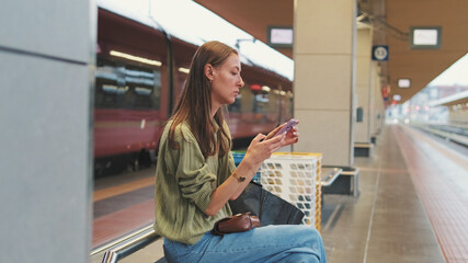 Young woman with brown hair, dressed in an olive green sweater, uses a mobile phone while sitting on a bench at a railway station