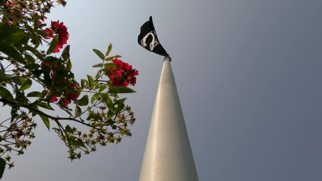 Metal flagpole with Prisoner of War (POW) flag waving against a clear sky. A flowering bush with reddish pink petals is in the forefront of view.