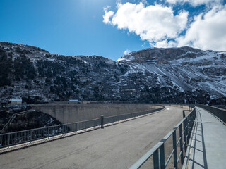 Valley of Lei. Swiss. Dam. Mountain Dam Under Blue Skies. A dam with a road atop, nestled between snow-dusted mountains under a clear blue sky.