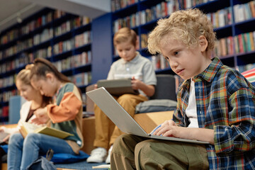 Side view portrait of blonde little boy using laptop in colorful school library with copy space