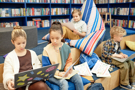 Portrait of two girls using laptop in modern library lounge with group of children in backgroundwith copy space