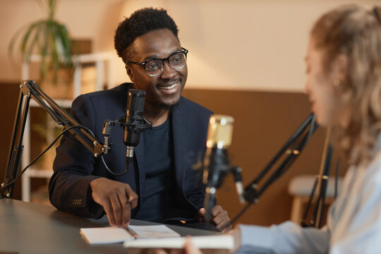 Medium shot of cheerful African American man wearing glasses and black suit talking to unrecognizable female guest sitting at table during podcast in studio