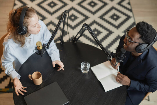 Top down view of young Caucasian woman and cheerful Black man holding interview sitting at black round table in podcast studio