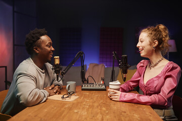 Side view of young African American man and trendy Caucasian girl having fun during podcast recording sitting at desk in studio with neon light