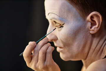 Side view closeup of adult artist doing mime makeup by mirror backstage preparing for performance,...