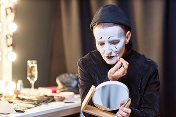 Dramatic portrait of female mime performer looking in mirror backstage and doing makeup preparing...