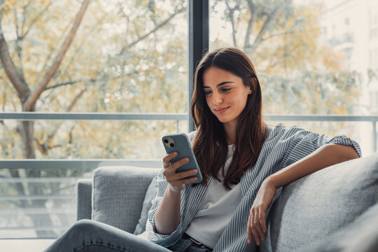 Happy pleasant millennial woman relaxing on comfortable couch, holding smartphone in hands. Smiling young lady chatting in social networks, watching funny videos, using mobile applications at home..