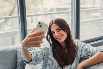 Happy pleasant millennial woman relaxing on comfortable couch, holding smartphone in hands. Smiling...
