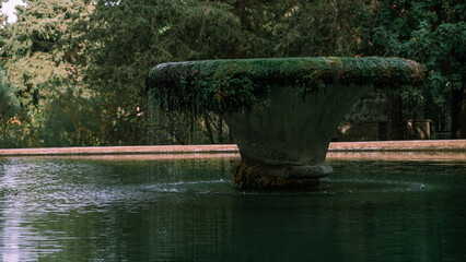 A large, mossy fountain sits in a pond. The water is calm and the surrounding area is lush and green