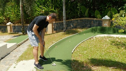 Active adult man playing mini golf on sunny day, enjoying outdoor leisure activities on well-maintained putting green. Outdoor sports and entertainment.