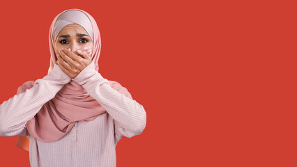 Frightened face. Omg panic. Shocking news. Scared speechless worried woman in hijab covering mouth isolated on red empty space background.