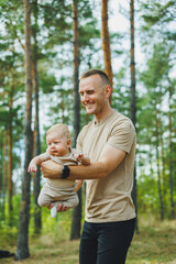 A young dad holds his little son in his arms while standing in the park. Dad is playing with his son