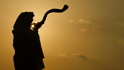 Silhouette of a Jewish man wearing a tallit and tefillin, blowing a long, curly shofar made from...
