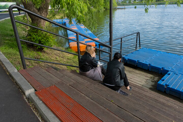 Two women sitting on the steps of a pier by a lake drinking coffee. Two friends having lunch by the...
