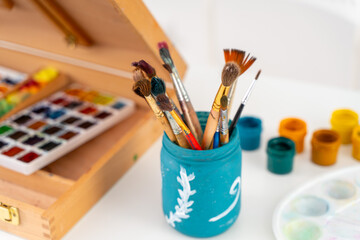 Artist's brushes in a turquoise jar, a malbert with paints and jars with acrylic paint on a white table. Concept for the development of children's creativity. Selected Focus