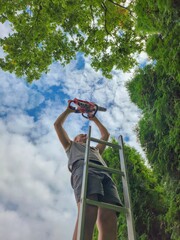 Man climbing a ladder with a chainsaw under the bright sky, surrounded by lush green trees in a forest on a sunny day, showing harmony between nature and human activity in a picturesque setting.