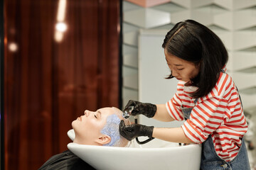 Side view portrait of young Asian hairstylist washing hair of young woman in salon sink and giving...
