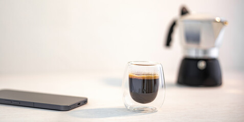 Espresso in a glass cup beside a smartphone, with a moka pot in the blurry background.