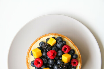 Cheesecake decorated with fresh berries and mango. On a light gray plate. White background. Back view. There is a copy space for your text.